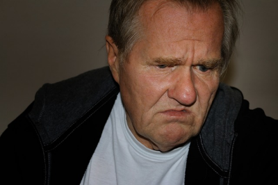 An older white man wearing a white shirt and black hooded sweatshirt looks down and to the right with an expression of resigned disappointment.